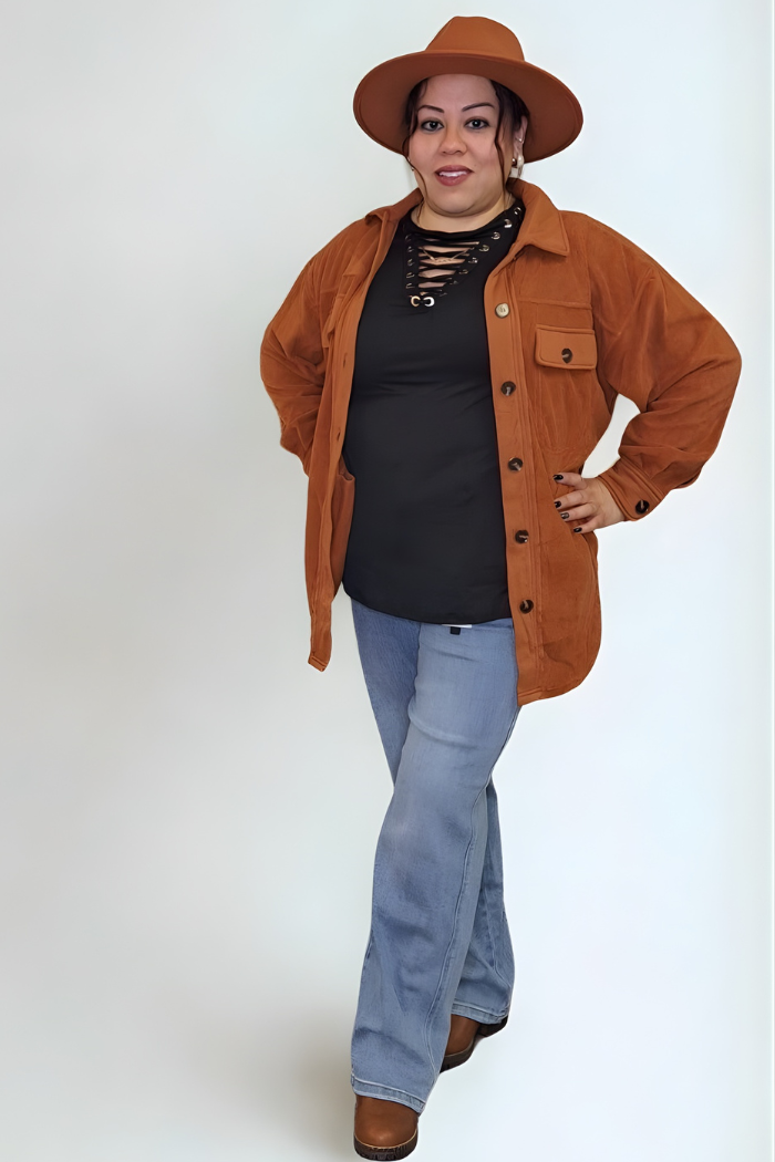 A woman wearing a black lace-up top, a rust-colored shacket, and light-wash flare jeans, with a rust fedora-style hat. She is standing with her hands on her hips and smiling at the camera.