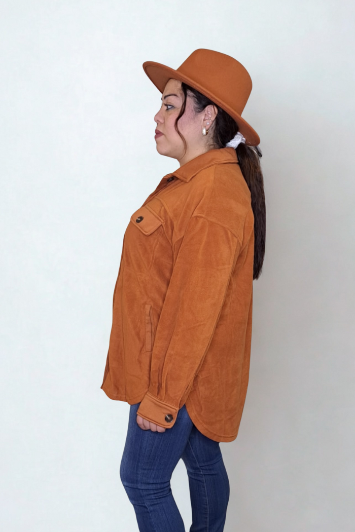 A woman in a rust-colored shacket and a matching hat is shown in a profile view, looking to the left. She is wearing blue jeans and has her hair in a ponytail. The shirt jacket has two chest pockets with buttons. 