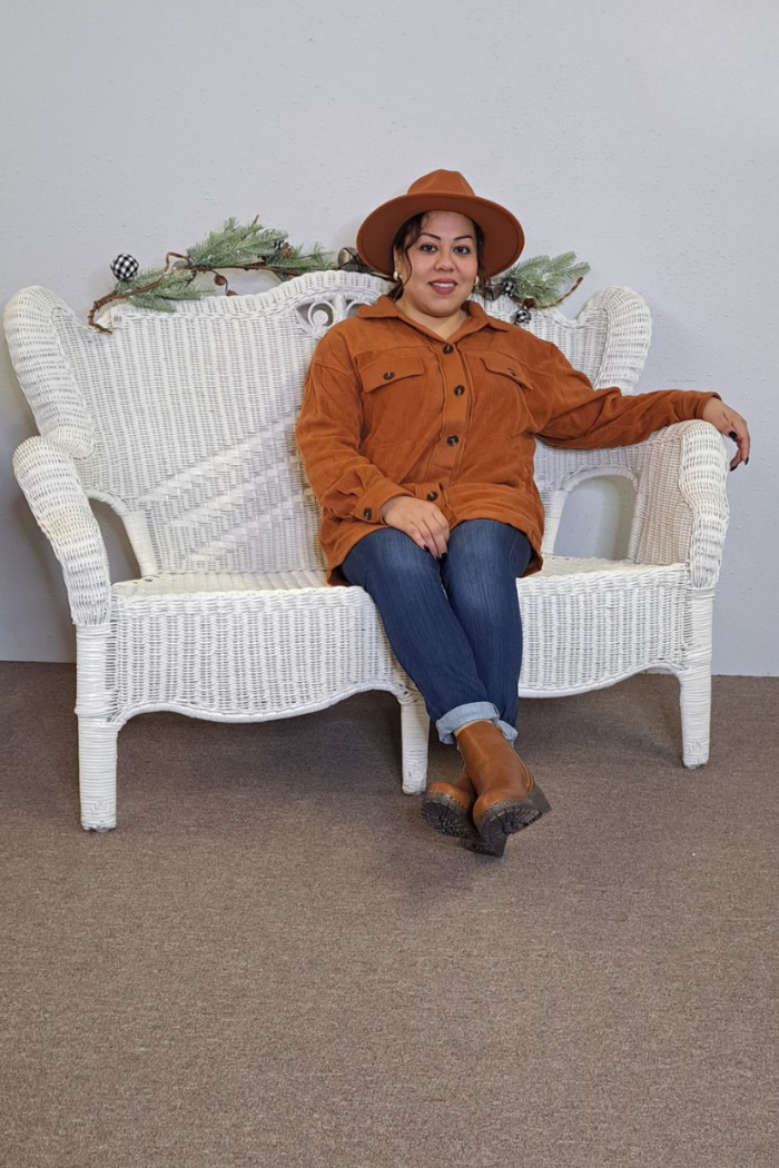 A woman wearing a rust shacket, a hat, and jeans is sitting on a white wicker bench with greenery draped over the back. She is smiling and looking at the camera. She is wearing brown boots.