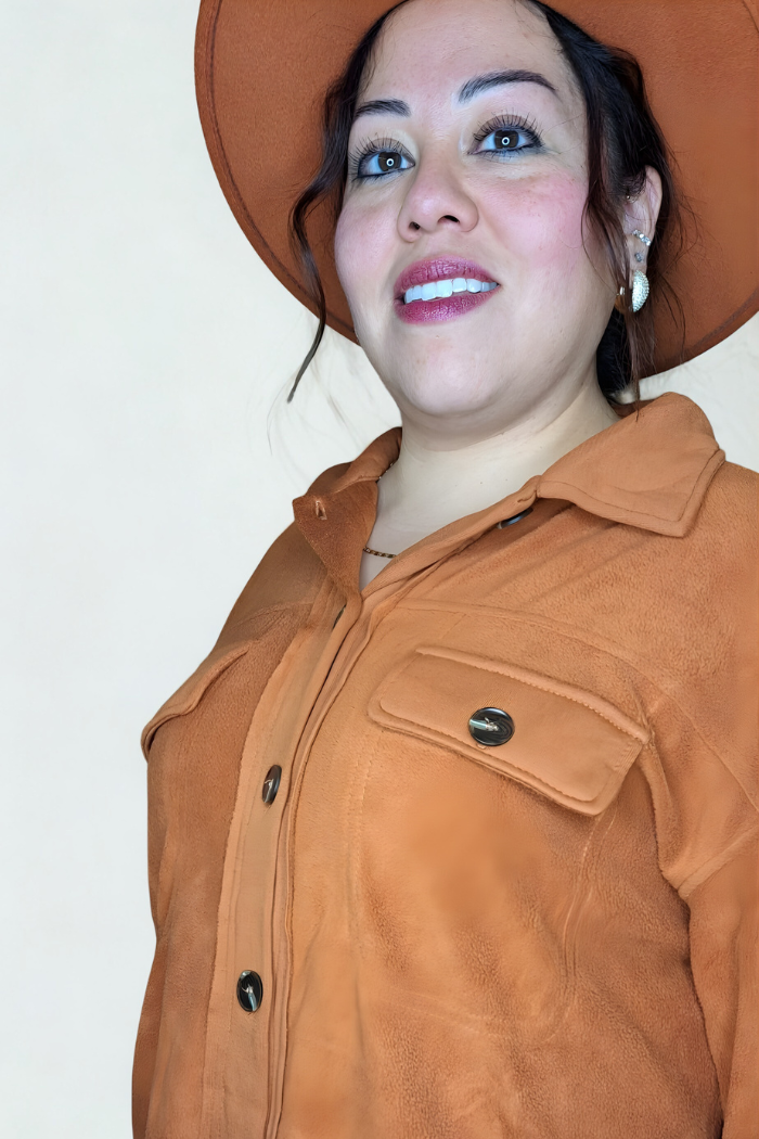 A woman wearing a rust button-down shacket and a matching hat, looking up and to the side with a soft smile.  She has dark, curly hair, brown eyes, and is smiling faintly with red lipstick. Her outfit features a collar and patch pockets on the chest with metallic buttons. She is also wearing earrings that appear to be white or pearl-colored.