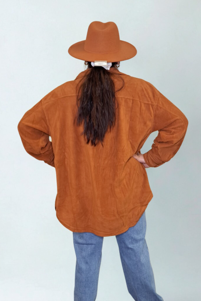 A person is shown from behind, wearing a rust-colored shacket, blue jeans, and a matching rust-colored hat. Their long brown hair is in a low ponytail secured with a white scrunchie. She is standing with their hands on their hips against a light gray background.