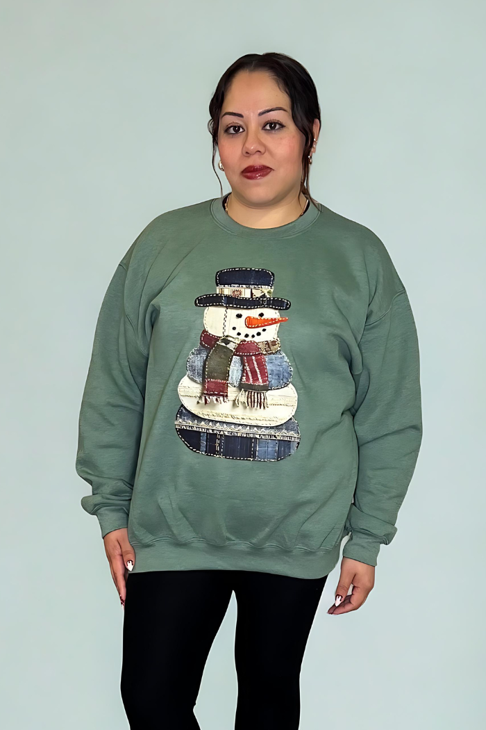 A woman with dark hair pulled back smiles softly at the camera, wearing the Snowman Patchwork Holiday Sweatshirt and black leggings against a plain background.