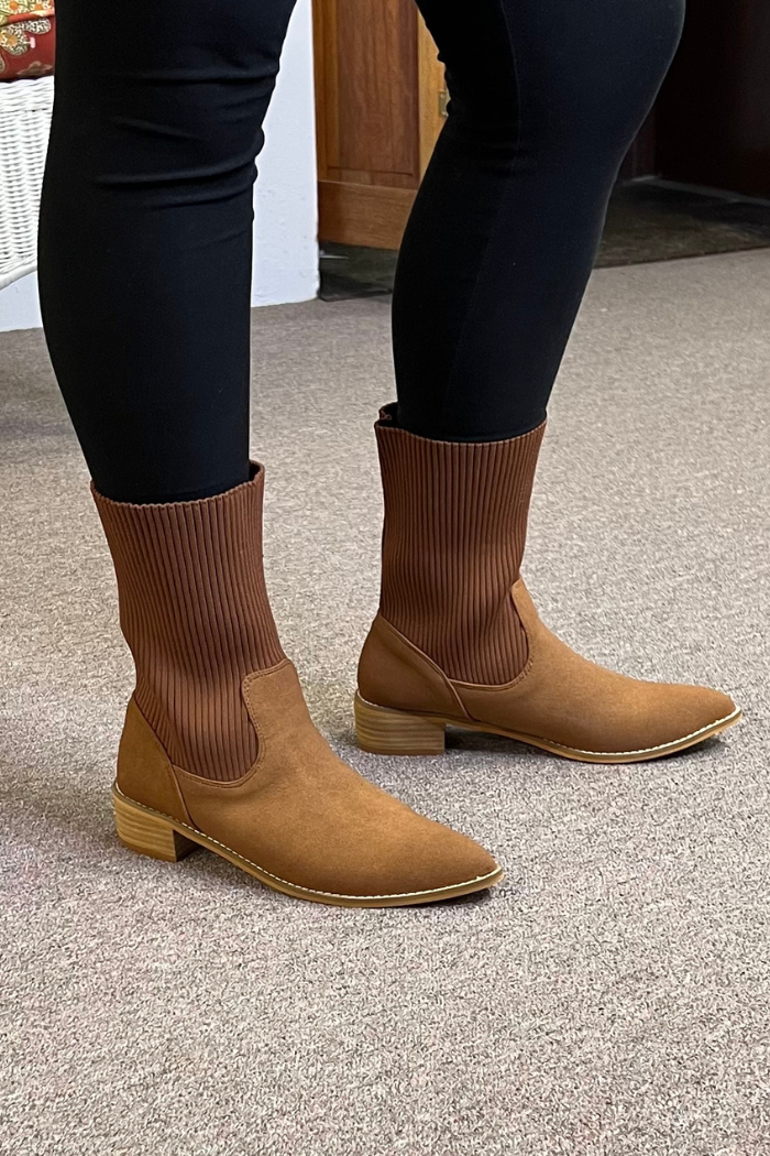 Someone wearing black leggings and Folded Knit Ankle Booties with a brown ribbed knit cuff and low wooden heel stands on a beige carpet indoors.