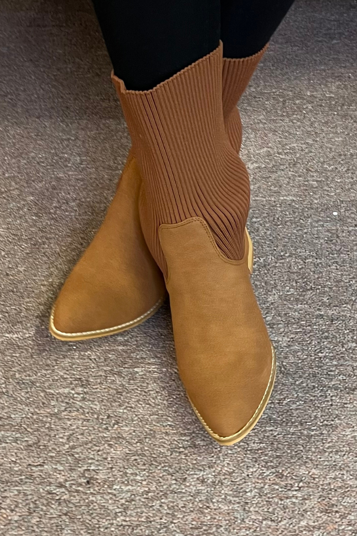 Someone stands on a brown carpeted floor wearing light brown Folded Knit Ankle Booties featuring pointed toes and a ribbed knit cuff.
