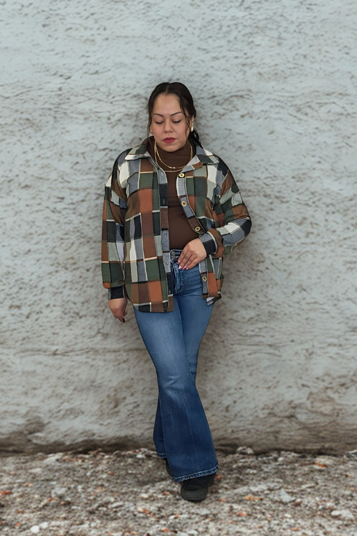 A woman stands before a textured light wall, wearing the Plaid Print Shirt Jacket in brown and green over a brown shirt, paired with blue flared jeans and black shoes. She gazes downward with one hand in her pocket.