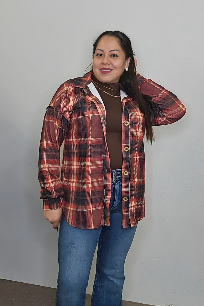 A woman smiles in a Plus Size Plaid Flannel Shacket over a brown turtleneck and blue jeans, touching her hair against a plain light wall—an ideal cozy fall look.