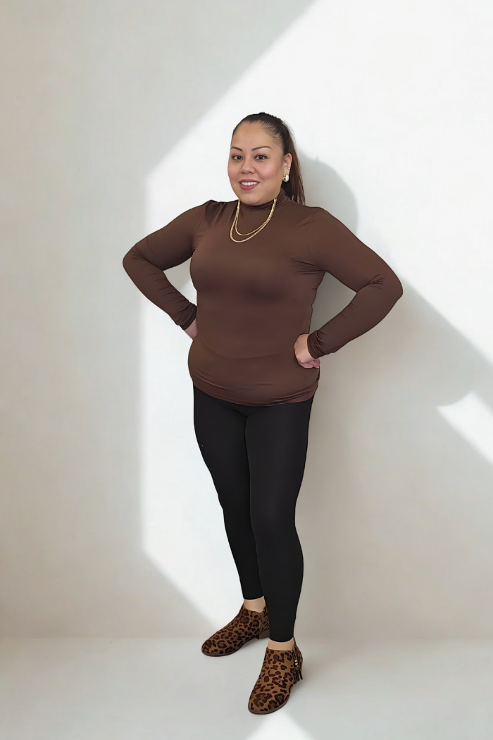 A woman smiles in sunlight against a white wall, wearing the Lightweight Mock Neck Shirt in brown with black leggings, gold jewelry, and leopard-print shoes—delivering an effortless take on women's workwear, hands on hips.