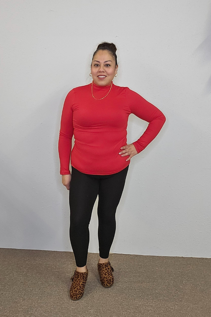 A woman stands against a light wall, smiling with one hand on her hip. She's wearing a red Lightweight Mock Neck Shirt styled as workwear, black leggings, leopard-print shoes, and has her hair tied up.