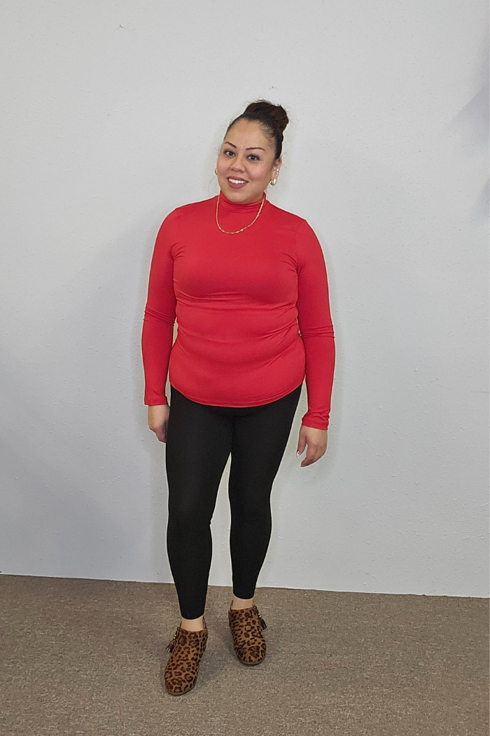 A woman smiles against a white wall, wearing the Lightweight Mock Neck Shirt as her long-sleeve top, styled with black leggings, a gold necklace, leopard print shoes, and her hair in a bun—a chic approach to women’s workwear.