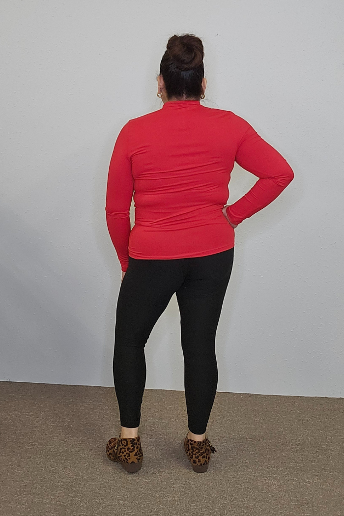A person with dark hair in a bun stands with their back to the camera, wearing a red Lightweight Mock Neck Shirt, black leggings, and leopard print shoes against a plain light wall and carpeted floor.