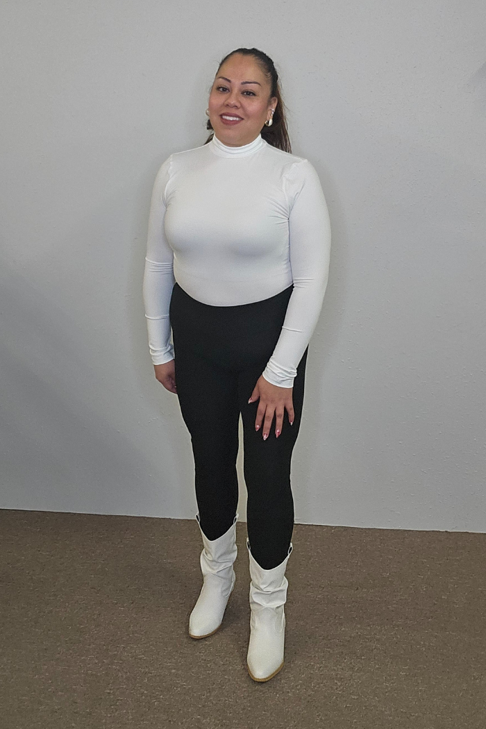A woman smiles against a plain light wall, wearing the Lightweight Mock Neck Shirt in white with long sleeves, black pants, and white boots—ideal for stylish women’s workwear. Her hair is tied back in a neat ponytail.
