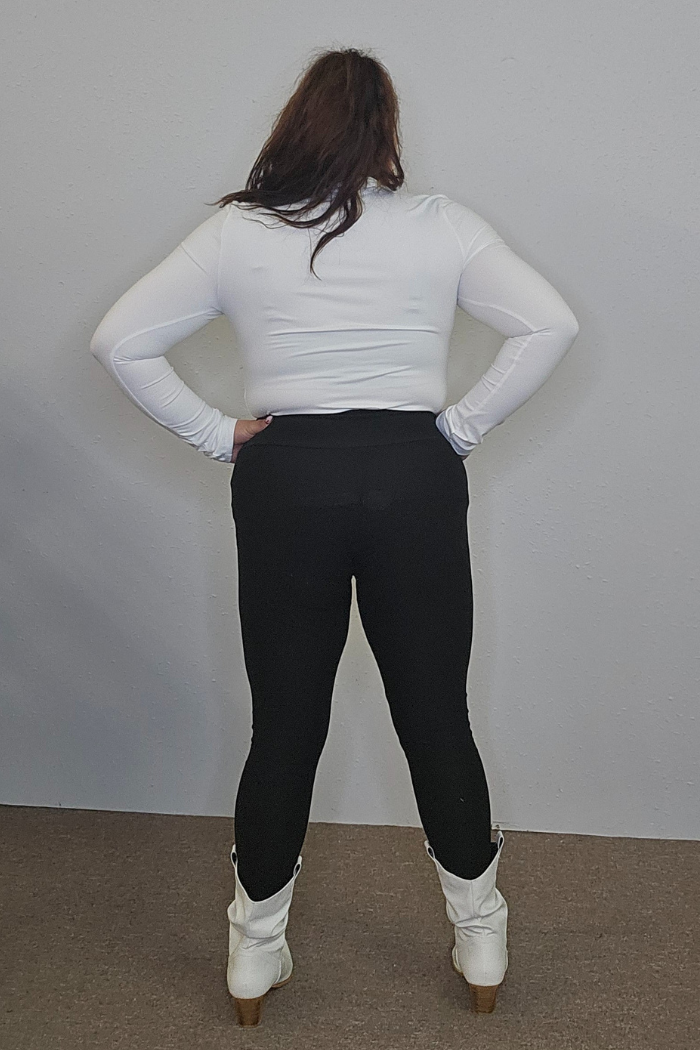 A person with long dark hair faces away from the camera, displaying chic women's workwear: a Lightweight Mock Neck Shirt paired with black pants and white heeled boots against a plain light-colored wall.