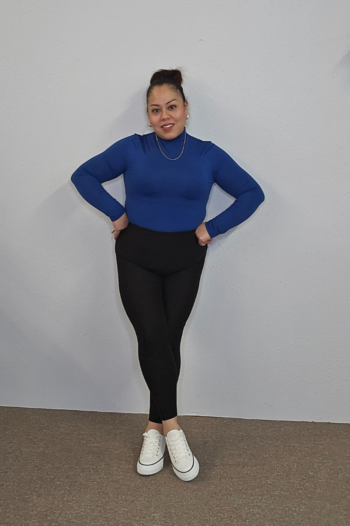 A woman stands against a light gray wall, smiling with her hands on her hips. She wears the Lightweight Mock Neck Shirt in blue, paired with black leggings and white sneakers. The carpeted floor is a neutral shade.