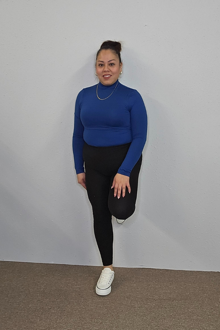 A woman in a long-sleeve blue Lightweight Mock Neck Shirt, black leggings, and white sneakers poses with one leg bent against a plain light gray wall—an ideal choice for chic women's workwear.