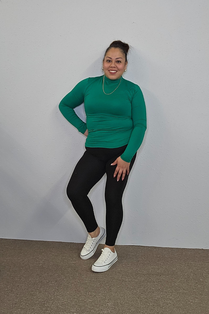 A woman with her hair in a bun poses confidently against a light gray wall, wearing the Lightweight Mock Neck Shirt in green, black leggings, and white sneakers—showcasing effortless women's workwear style.