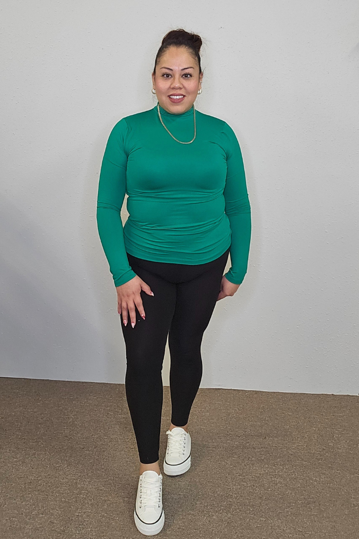 A woman stands indoors by a plain light wall, smiling in the Lightweight Mock Neck Shirt styled as women's workwear, paired with black leggings, white sneakers, and a gold necklace. Her hair is tied up for a polished look.