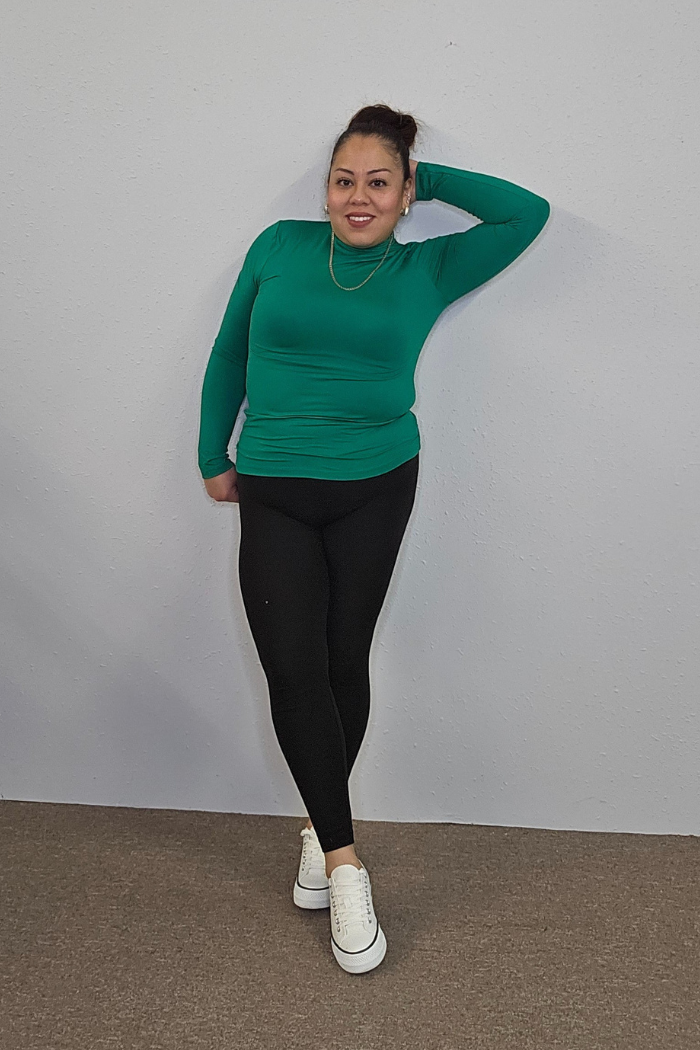 A woman poses against a light gray wall in the Lightweight Mock Neck Shirt and black leggings, styled with white sneakers and a gold necklace for an effortlessly chic workwear look. She smiles, one hand on her hip, the other behind her head.