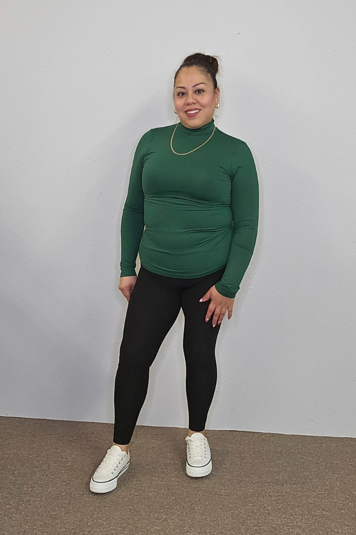 A woman smiles indoors against a plain wall, wearing a green Lightweight Mock Neck Shirt, black leggings, white sneakers, gold hoop earrings, and a gold necklace—an easy, stylish workwear look with hair in a bun.