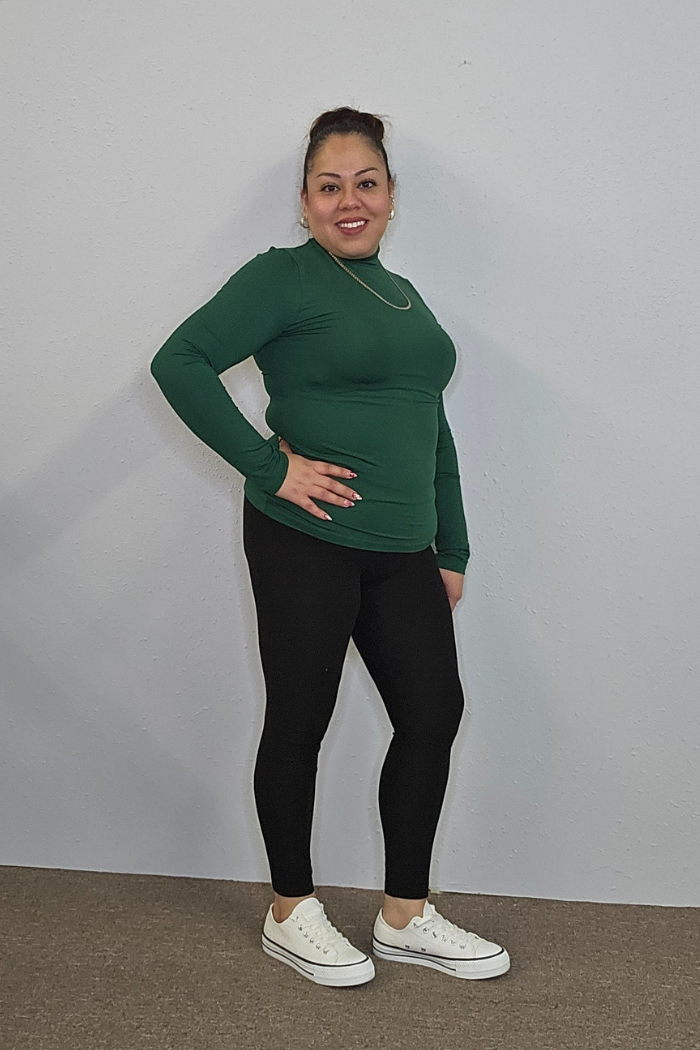 A woman smiles against a light gray wall, wearing the Lightweight Mock Neck Shirt in green with black leggings and white sneakers. Her hair is tied up, and she stands confidently with one hand on her hip.
