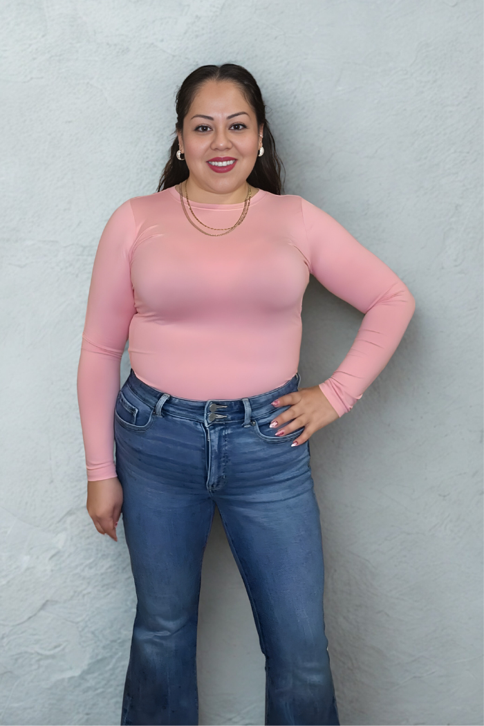 A woman with long dark hair, wearing a Brushed Microfiber Long Sleeve Top in soft pink, blue jeans, and gold necklaces, smiles while standing against a light gray textured wall.