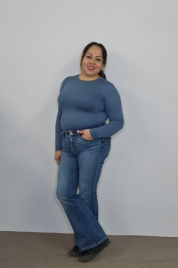 A woman with long dark hair smiles at the camera against a plain light wall, wearing a buttery soft Brushed Microfiber Long Sleeve Top in blue, blue jeans, and black shoes, with one hand in her pocket.