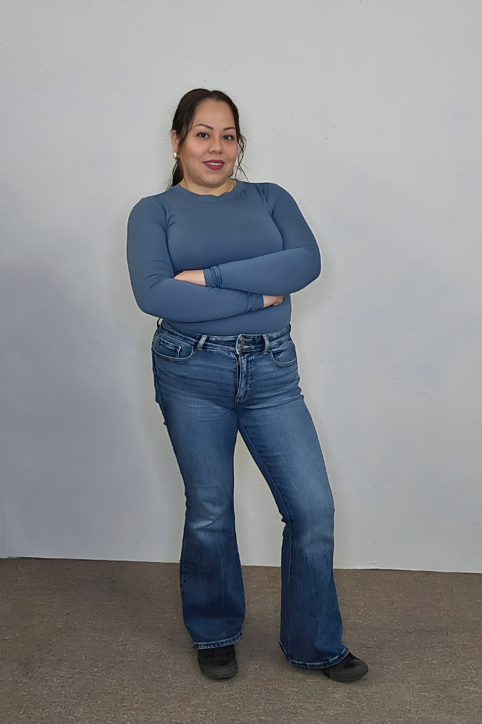 A woman with long dark hair stands with arms crossed, wearing the Brushed Microfiber Long Sleeve Top, blue jeans, and black shoes in front of a plain light-colored wall.