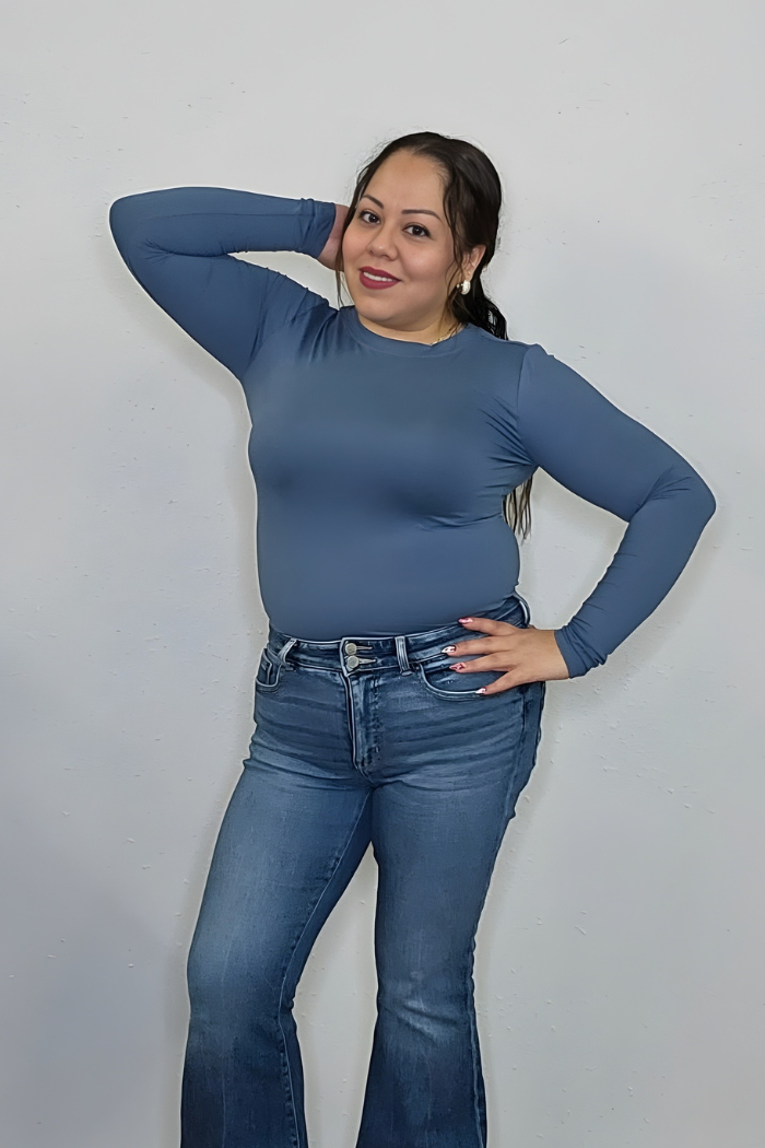 Woman with long dark hair smiles at the camera, posing in a Brushed Microfiber Long Sleeve Top and blue jeans against a plain light-colored wall, with one hand on her hip and the other behind her head.