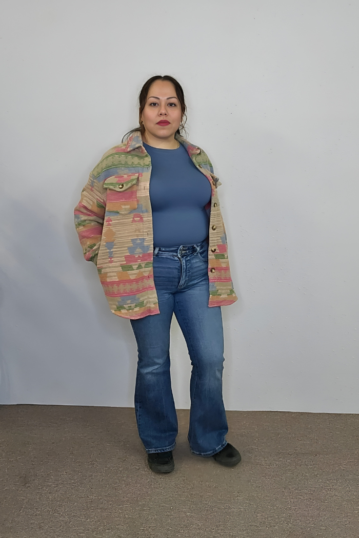 A woman stands against a plain wall, wearing a patterned beige jacket, Brushed Microfiber Long Sleeve Top in soft blue, blue jeans, and black shoes. She has her left hand in her pocket and looks directly at the camera.