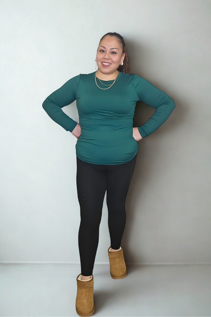 A woman stands against a plain light background, smiling with her hands on her hips. She wears a teal Brushed Microfiber Long Sleeve Top, black leggings, tan slip-on shoes, and gold jewelry.