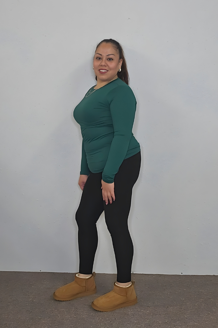 A woman with long dark hair in a ponytail stands against a plain light wall, smiling at the camera in a green Brushed Microfiber Long Sleeve Top, black leggings, and tan boots.