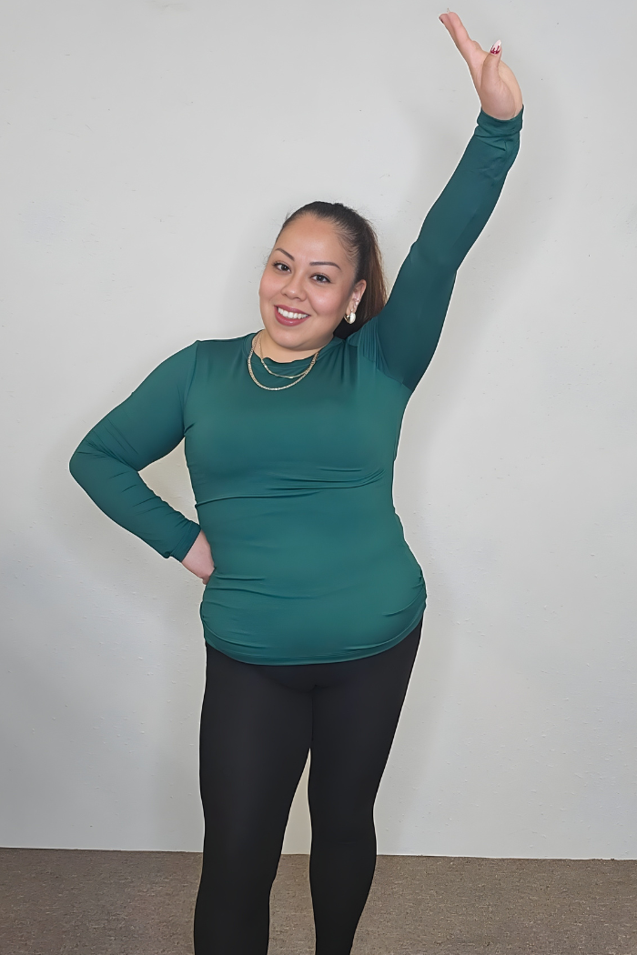 A woman wearing the Brushed Microfiber Long Sleeve Top in green and black leggings stands indoors, smiling, with one hand on her hip and the other arm raised against a plain light-colored wall.