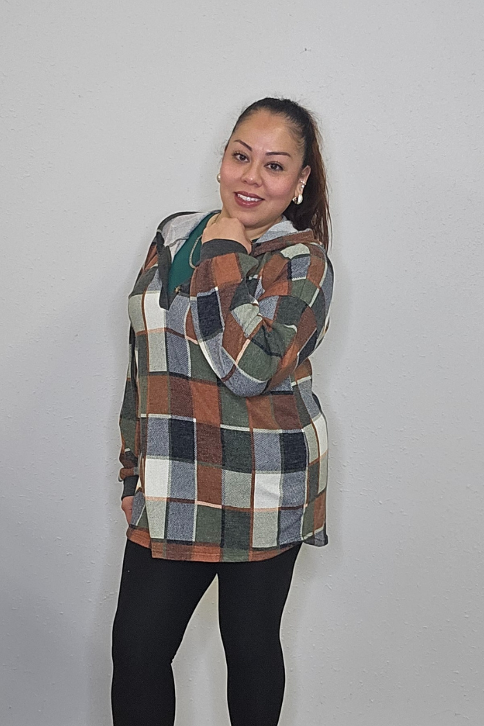 A woman with a ponytail smiles at the camera in a green and brown plaid shirt, green top, and black leggings against a light gray wall, showing off her cozy style, which could be completed with the Plus Multi-Color Square Hoodie.