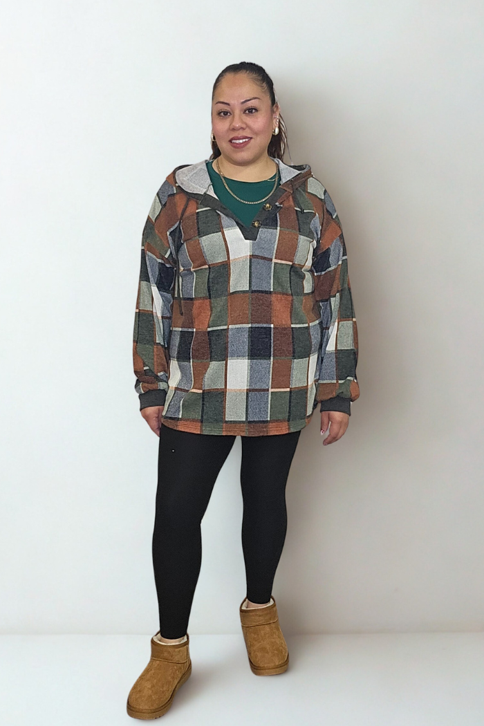A woman stands against a white background, wearing a green shirt, black leggings, tan slip-on shoes, and the Plus Multi-Color Square Hoodie featuring brown and green plaid. Her hair is tied back and she smiles at the camera.