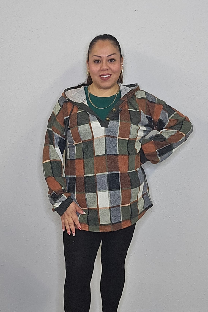 A woman stands smiling in front of a light gray wall, wearing a green shirt, black leggings, and the Plus Multi-Color Square Hoodie featuring plaid shades of brown, orange, gray, and green. One hand is on her hip.