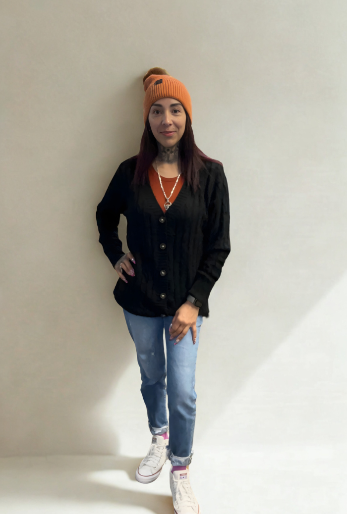 A woman with straight dark hair stands against a plain light background, wearing an orange beanie, orange shirt, blue jeans, white sneakers, and the Oversized Knit Button Cardigan. She looks at the camera with a relaxed posture.