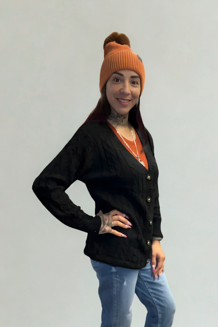 A woman wearing an orange beanie, blue jeans, necklaces, and an Oversized Knit Button Cardigan stands with one hand on her hip, smiling at the camera against a plain light background.