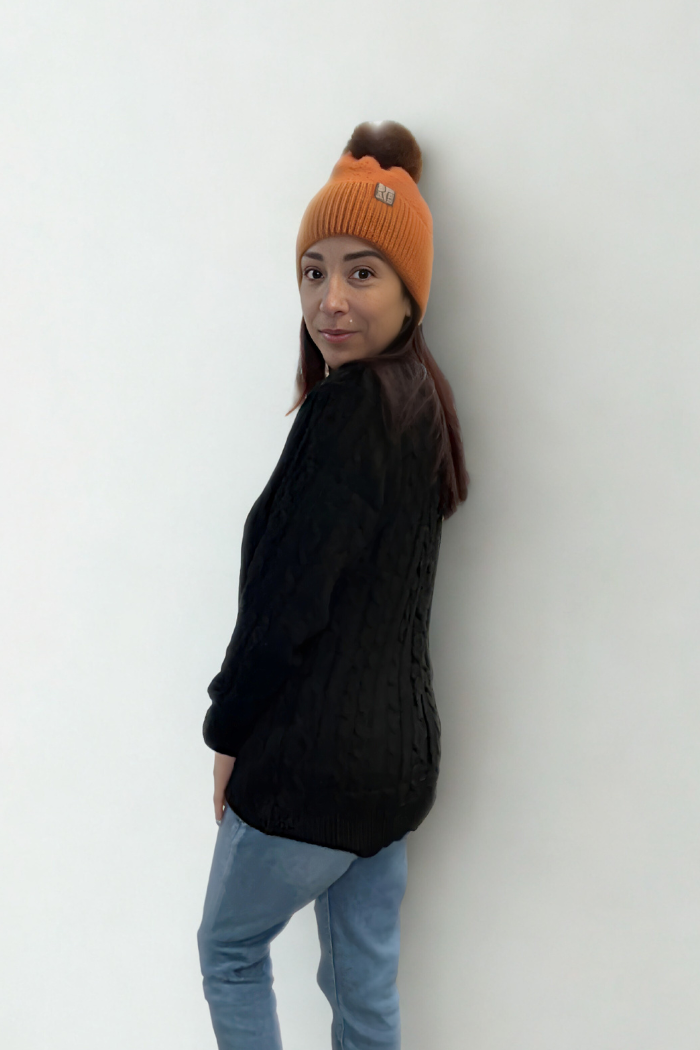 A person with long dark hair stands sideways and looks at the camera against a plain white background, wearing an Oversized Knit Button Cardigan, blue jeans, and an orange knit beanie.