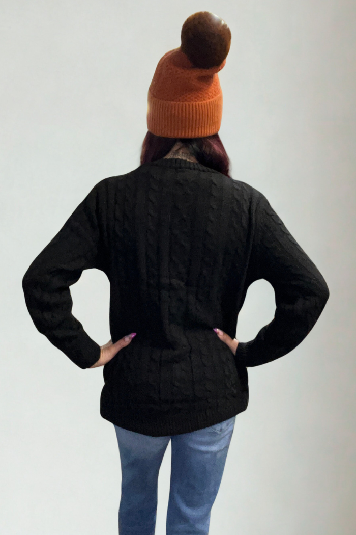 A person stands facing away, wearing an oversized knit button cardigan, blue jeans, and an orange pom-pom beanie. Their hands are on their hips against a plain light background.