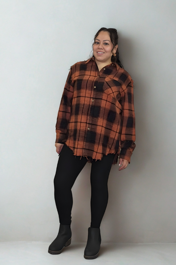 A woman stands against a light gray wall, smiling, wearing the Mineral Washed Plaid Shacket in brown and black plaid with black leggings and dark ankle boots. Her dark hair is tied back, and she accessorizes with large hoop earrings.