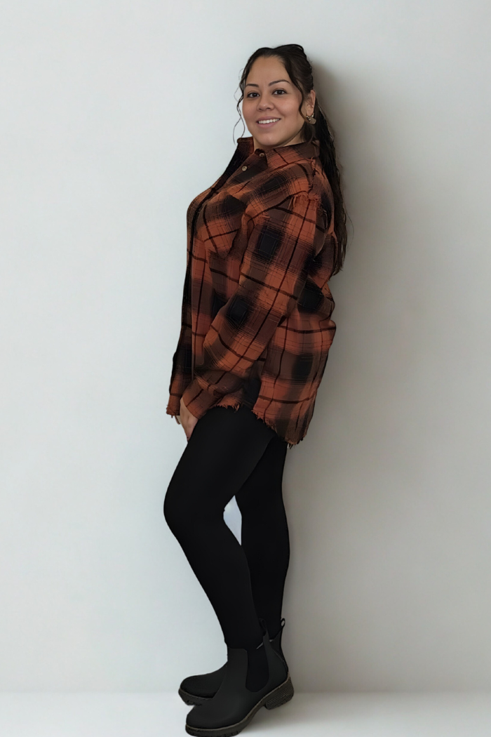 A woman with her long hair in a ponytail smiles while standing sideways against a plain light wall. She is wearing the Mineral Washed Plaid Shacket, paired with black leggings and black ankle boots.