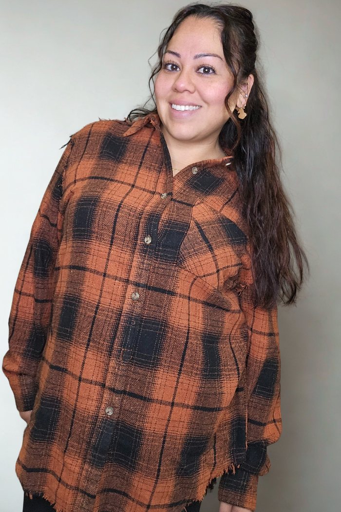 A woman with long, dark hair smiles at the camera wearing the Mineral Washed Plaid Shacket in orange and black. The plain, light background draws attention to her cozy, oversized style.