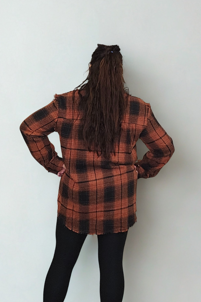 A person with long, wavy brown hair stands facing a plain white wall, wearing the Mineral Washed Plaid Shacket in orange and black plaid and black leggings, hands on hips.