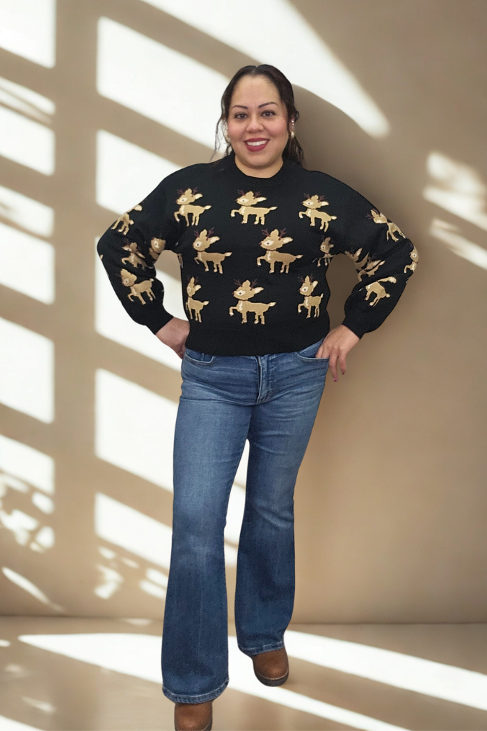 A woman stands smiling with hands on hips, wearing the Slouchy Reindeer Pullover Sweater featuring tan reindeer designs, paired with blue flared jeans and brown boots. Sunlight and window shadows cast patterns on the beige wall behind her.