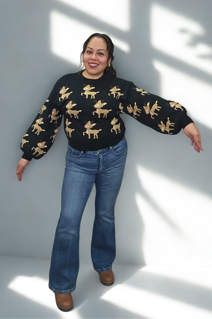 A smiling woman stands in a sunlit room, arms outstretched, wearing the Slouchy Reindeer Pullover Sweater with blue jeans and brown shoes. Shadows fall on the light gray wall behind her.