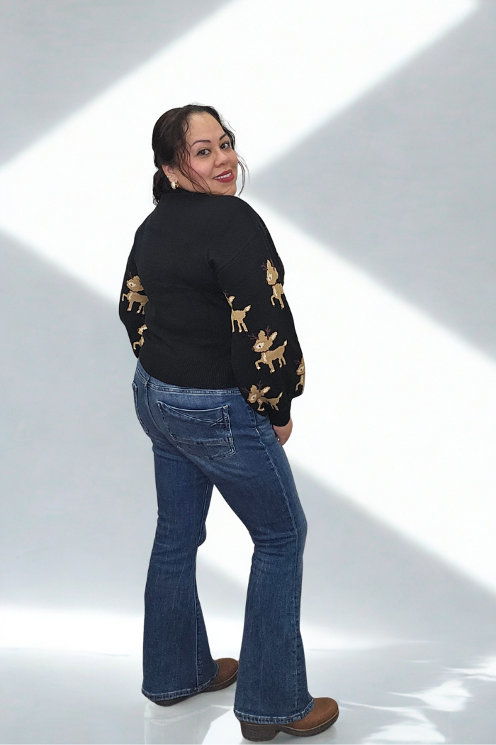 A woman with dark hair in a ponytail smiles over her shoulder, wearing the Slouchy Reindeer Pullover Sweater, blue jeans, and brown shoes. She stands against a light background with diagonal white lines.