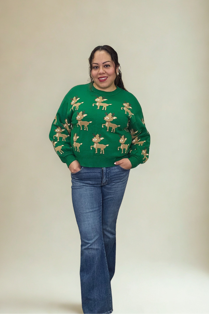 A woman smiles with her hands in the pockets of her jeans, wearing the green Slouchy Reindeer Pullover Sweater. The background is plain and light-colored.