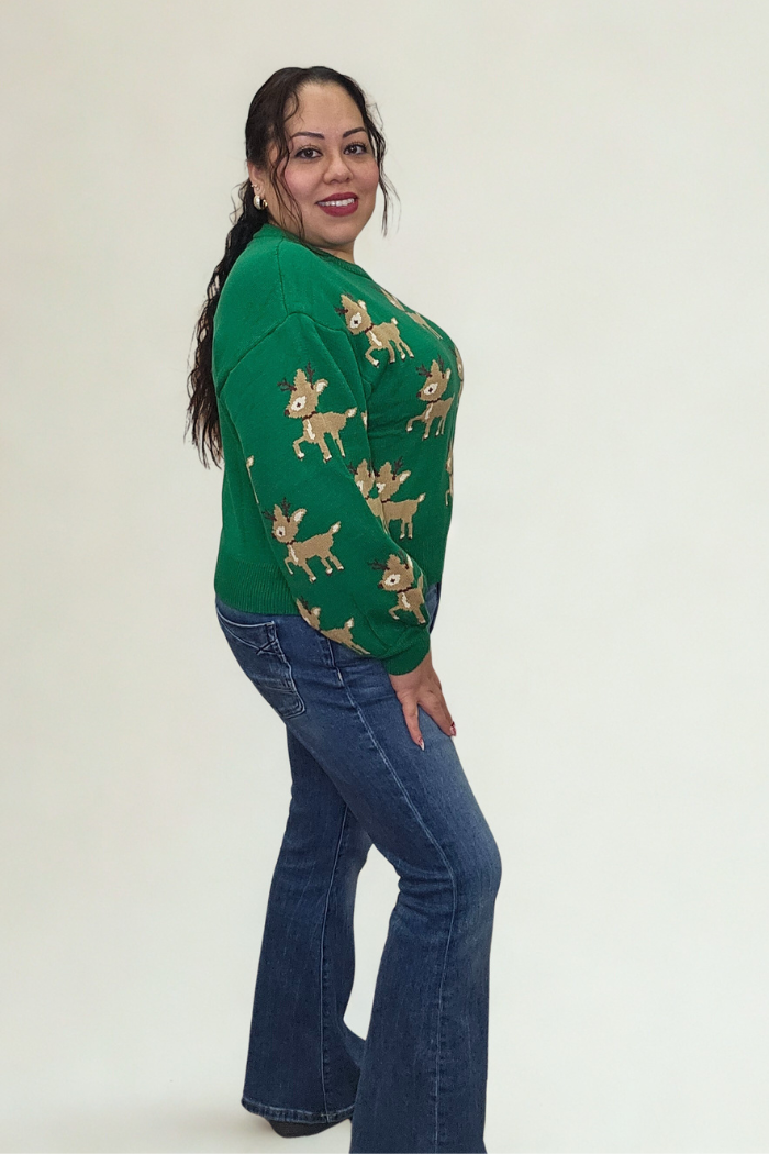 A woman with long dark hair smiles against a white background, wearing the green Slouchy Reindeer Pullover Sweater and blue jeans—an ideal festive holiday sweater.