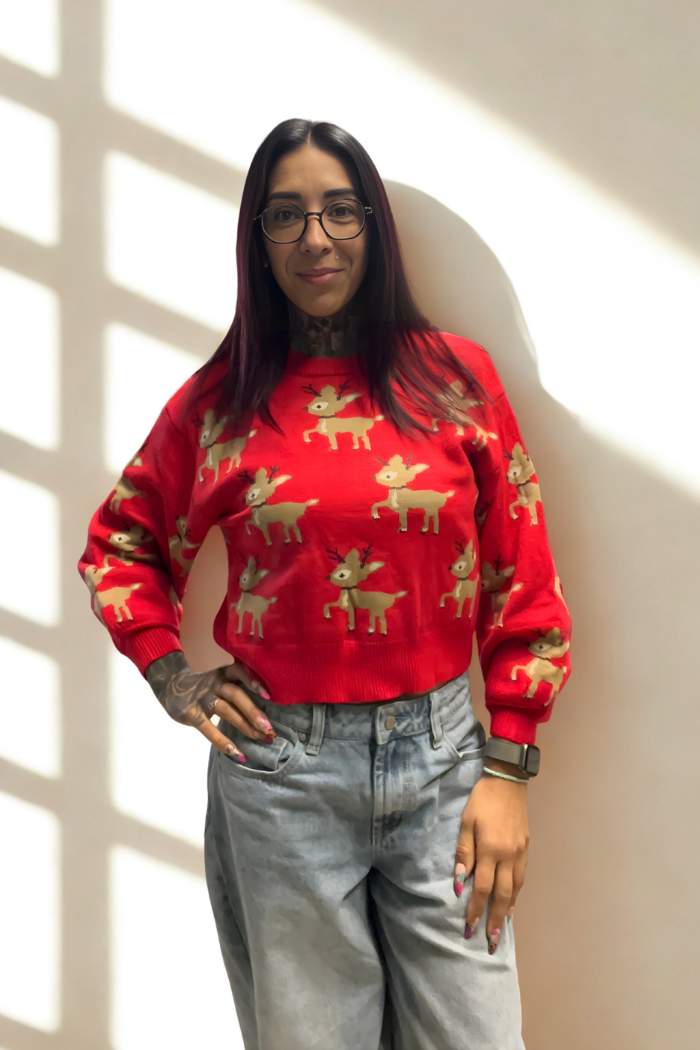 A woman with long dark hair and glasses wears the Slouchy Reindeer Pullover Sweater in red and light blue jeans, standing against a pale wall with window pane shadows.