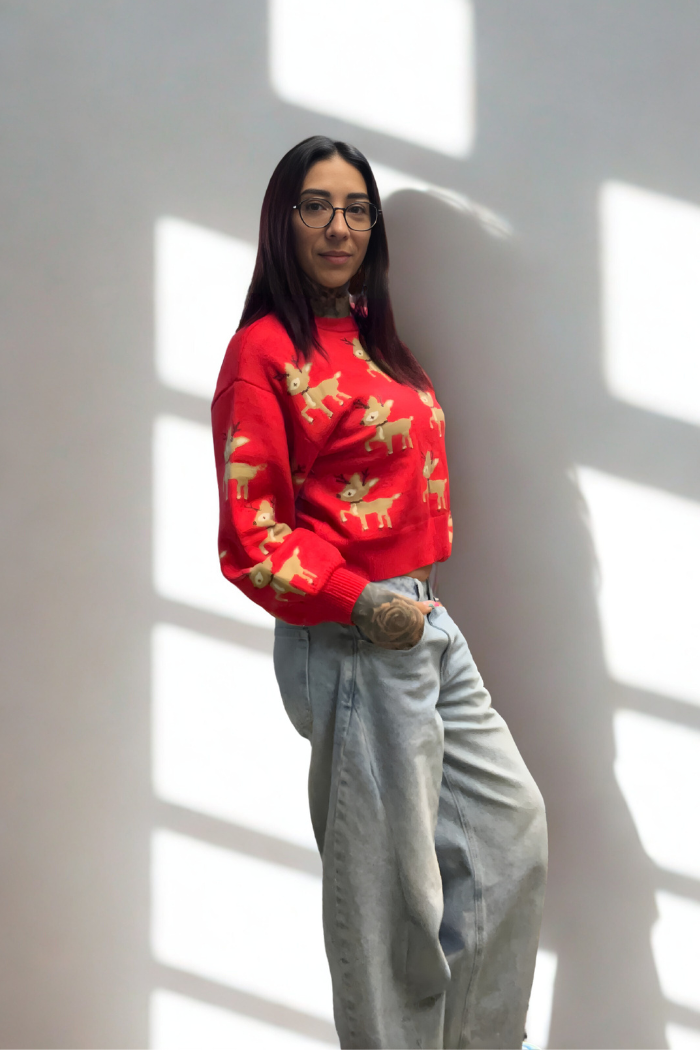 A woman with long dark hair and glasses stands against a light wall with window shadows, wearing the Slouchy Reindeer Pullover Sweater and light blue jeans, posing casually with one hand in her pocket and a relaxed expression.