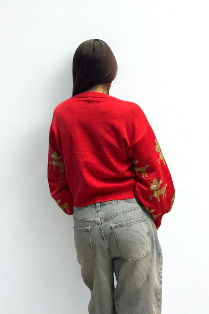 A person faces away from the camera, wearing a Slouchy Reindeer Pullover Sweater with festive sleeve designs and light gray, high-waisted jeans against a plain white background.